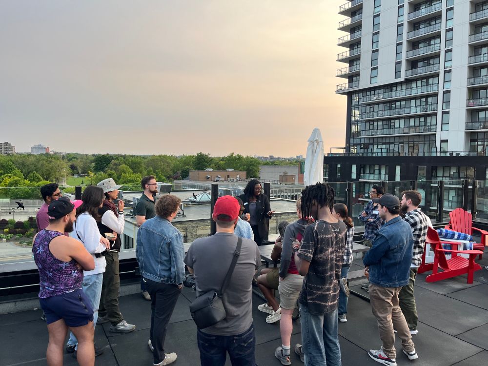 A candid rooftop shot of Colleen James speaking to a semicircle of attendees during an outdoor evening gathering. People are attentively listening, and a tall apartment building and treetops are visible in the background.