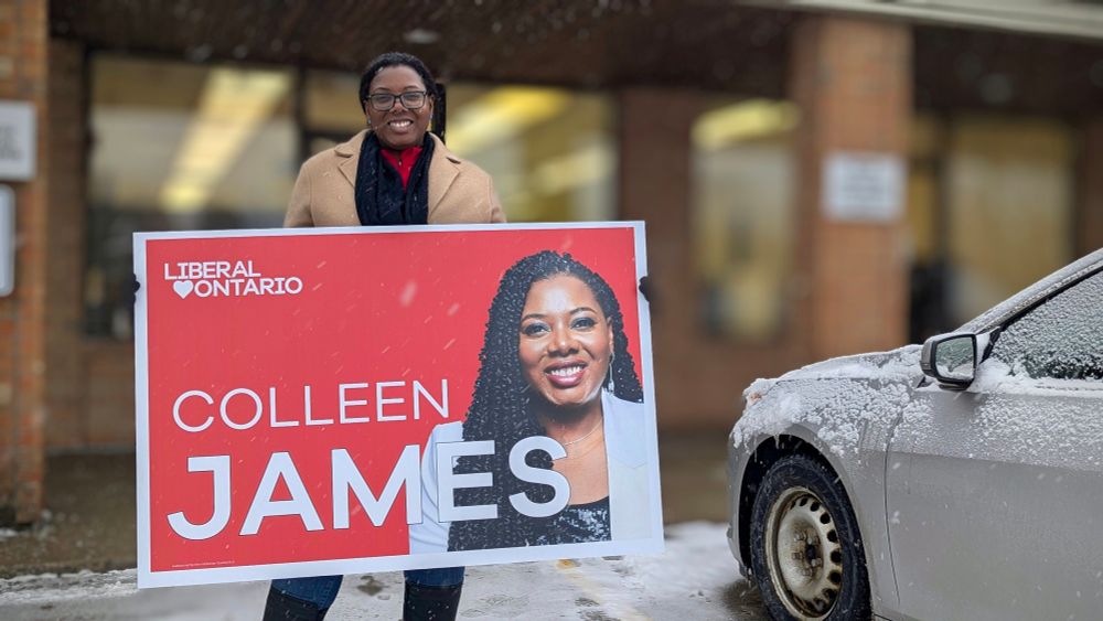 Colleen James wearing a brown tan coat holding a large Colleen James sign. The sign is red with white letters that say Colleen James with an image of Colleen on the right. 