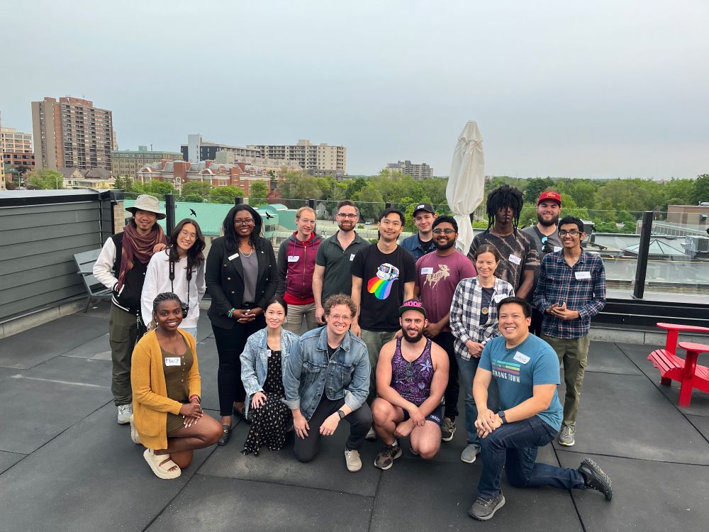 A group photo of around 20 people smiling together on a rooftop patio with a city skyline and trees in the background. Everyone is casually dressed and gathered after a StrongTowns WR event.