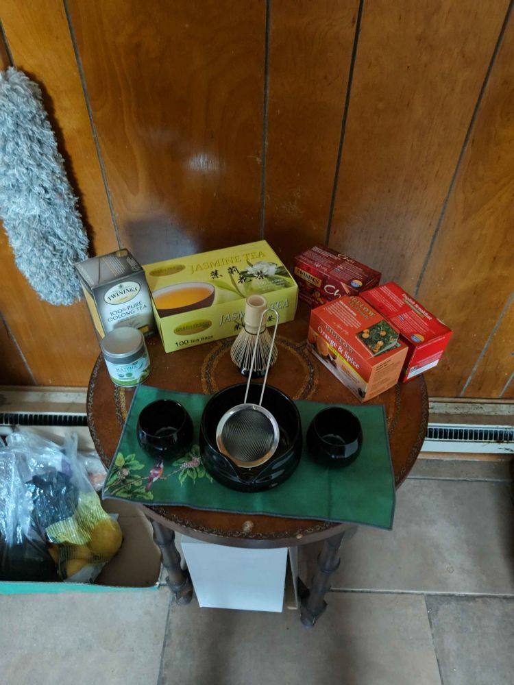 This image shows a tea preparation setup on a round wooden table with wood-paneled walls in the background. On the table is a green placemat with a traditional tea service arrangement including two dark tea cups, a central bowl with a wire whisk (likely for matcha preparation), and a fine mesh strainer. Behind the tea service are several boxes of tea including Twinings loose leaf oolong tea, jasmine tea (100 tea bags), and what appears to be chai tea boxes. There's also a small jar and other tea accessories. To the left side, there are some bags and a gray fuzzy item (possibly a cleaning tool or decorative object) visible. The setup suggests someone preparing for a traditional tea ceremony or tea tasting session.
