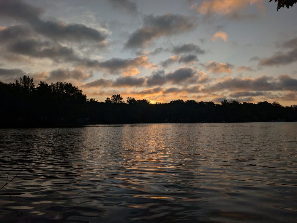 el amanecer sobre un lago. está un poco oscuro pero se puede ver el sol saliendo sobre los árboles y el cielo es azul, amarillo y naranja.