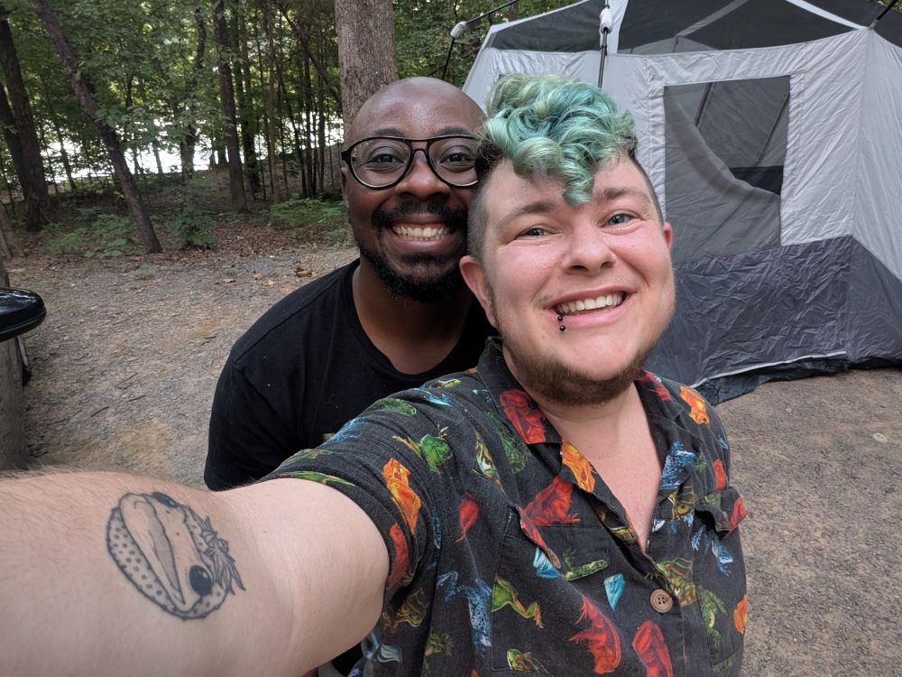 dos personas sonriendo a la cámara, de pie frente a una tienda de campaña y el bosque.