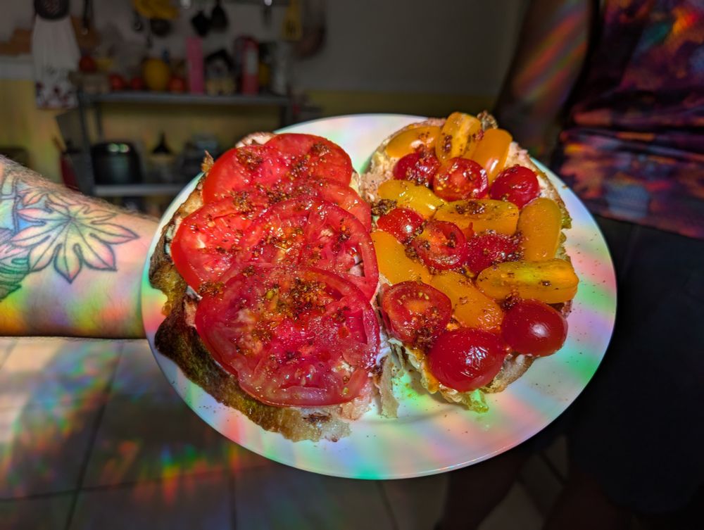 Un plato que contiene dos piezas de pan cubiertas de tomates. Una lleva tomates en rodajas y la otra, tomates cherry y uva. Los dos están rociados con aceite de chile. Los tomates son de un rojo intenso y amarillo.