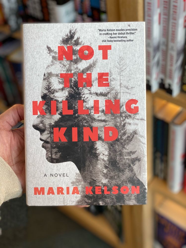 Holding a hardcover book with a cover of a silhouette of a woman’s profile with forest trees superimposed on it, with bookstore shelves in the background 