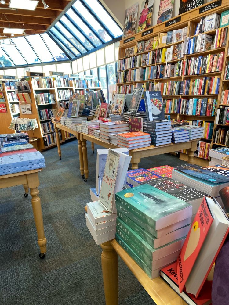 Bookstore with books displayed on tables among rows of bookshelves and large windows that extend to part of the ceiling in the background 