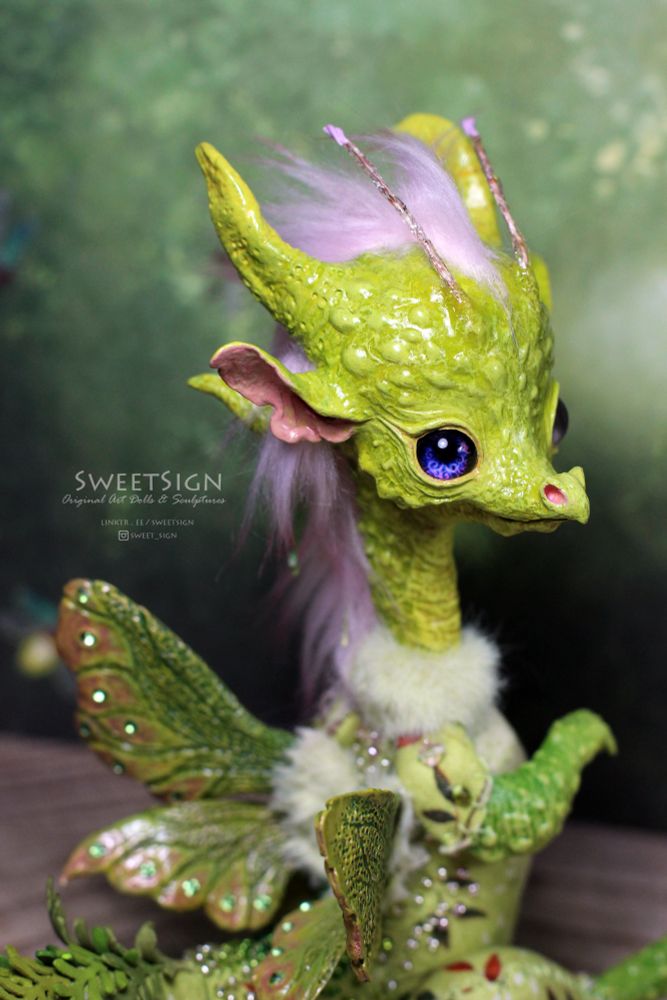 A light green longneck dragon with fairy wings and purple eyes, plants and moss overgrown ita tail. The creature is original miniature art doll I’ve made of airdry clay, acrylic paints, fabrics, faux fur and some other art supplies.