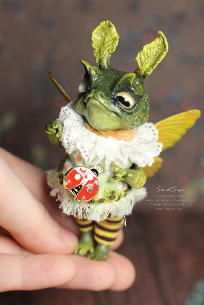 a teeny tiny original fantasy art doll, its head and legs sculpted of airdry clay, its body and outfit sewn of some fabrics and frills. The doll is Camilla the fairy frog, she has butterfly wings and moth' fluffy antennas, wears striped leggins and a dress with camomile print on green field, a ladybug pendant attached in the middle, and looks at you with a tint of smile. Such a capricious miniature lady she is.