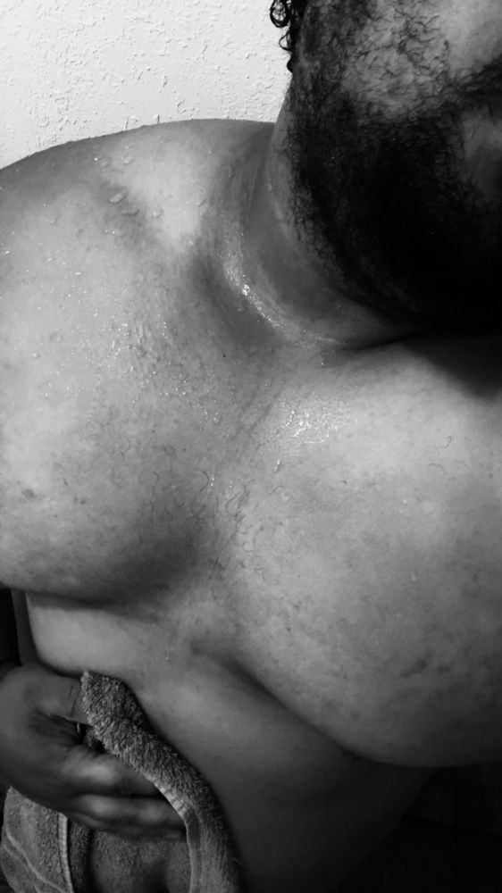 Picture Black and white color scheme. Photographed is Sunny post shower holding his towel over their stomach with one hand. Water drips from his chest and shoulders as his pecs and traps are being show off.