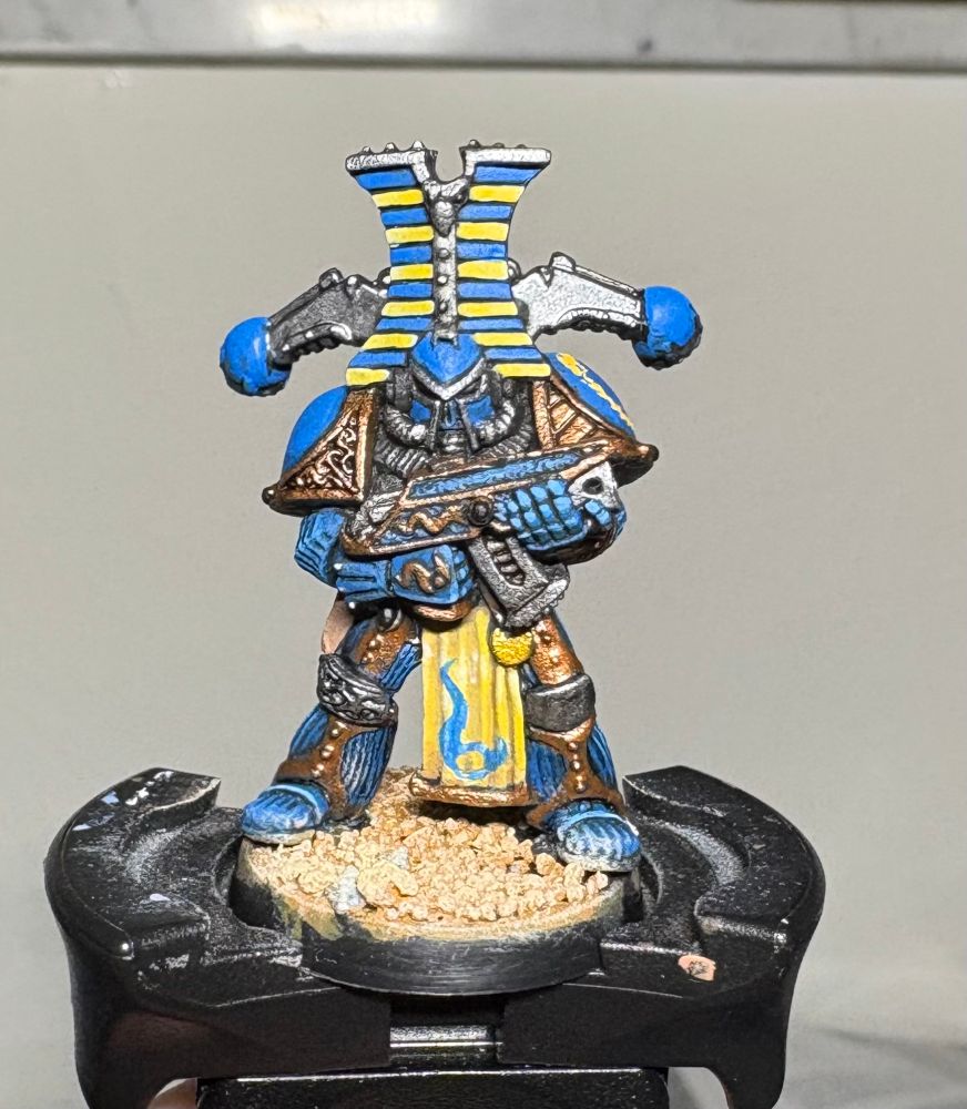 A rogue trader-era thousand sons chaos space marine mini for warhammer 40k. It is painted blue with steel and copper metal accents and yellow trim. The loin cloth has been painted yellow with a blue icon of Tzeentch freehand, after a reference image I found.