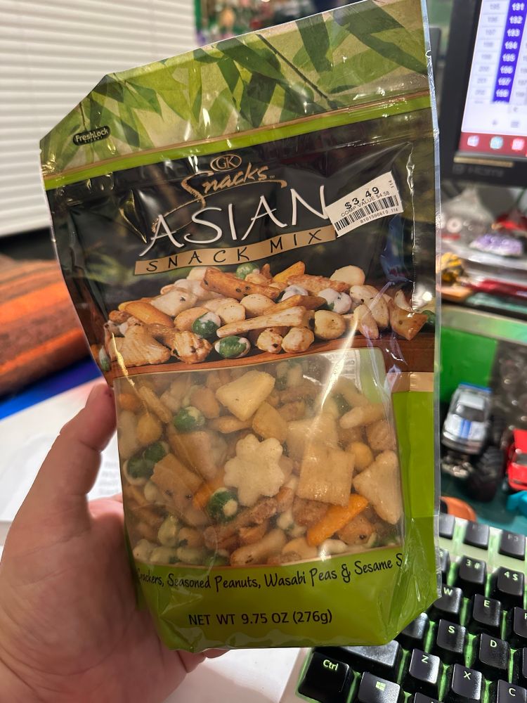 My hand, holding a big bag of GK Snacks Asian Snack Mix. Rice crackers of various types. A little wasabi kick but not so much it blows your sinus system out. This is the last bag from the several bags I bought before my local Big Lots stores went under. Not sure where to find them reliably now. That said, due to an upcoming medical procedure, I can’t eat stuff like this anymore, anyway. (Still want to find them for my wife though!)