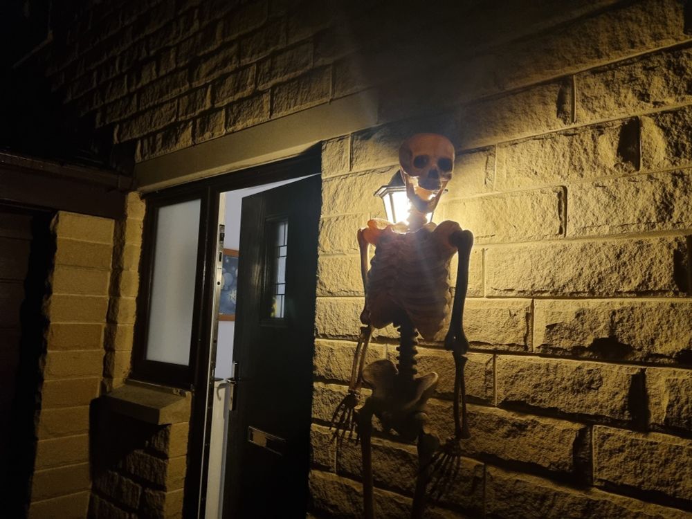 Photograph of a plastic life-sized skeleton Halloween decoration hanging up in front of a lamp outside the house next to the front door