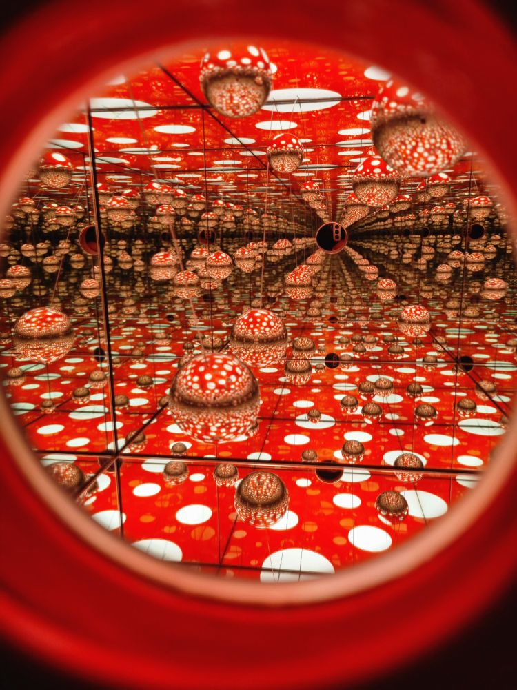 Photograph taken from the viewing hole of a smaller exhibition piece. When looking inside, there's an array of mirrors reflecting the image of several silver-coloured metal balls on top of a red surface with white polka dots. The reflections cause an optical illusion, making the overall view seem like it goes on forever in every direction, but is really just a small cube within a slightly bigger sphere. Elsewhere in the exhibit, there was a room that you could walk into that had exactly the same look and shape to it as this miniature version, making it feel like you had now entered the small world you saw here earlier