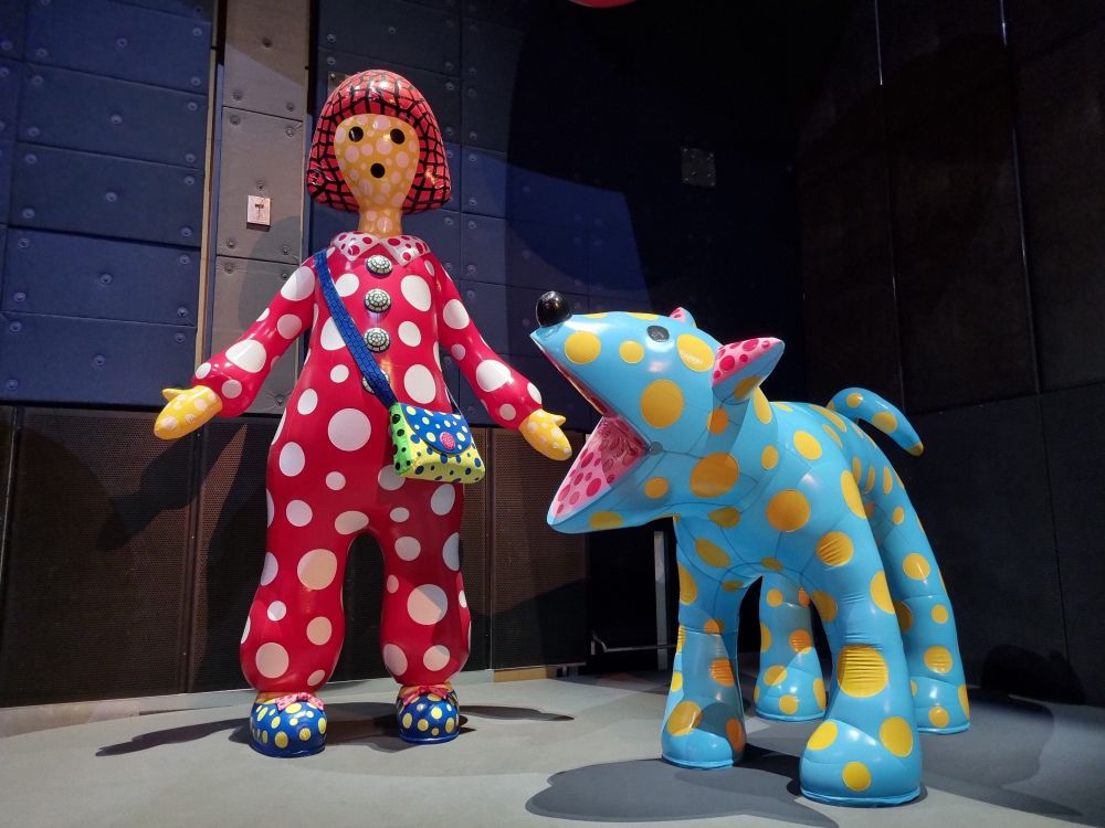 Photograph of a large inflated rubber girl wearing pink clothes with white polka dots, blue shoes with yellow polka dots. She has yellow skin with white polka dots and black hair with pink polka dots. She also has a purse that's green with black polka dots on the sides, yellow with blue polka dots in the middle and blue with yellow polka dots for the cover. Next to her is a large inflated rubber dog, who's light blue with yellow polka dots. The dog's mouth is open, as if barking, revealing a pink mouth with red polka dots, which matches its inner-ears