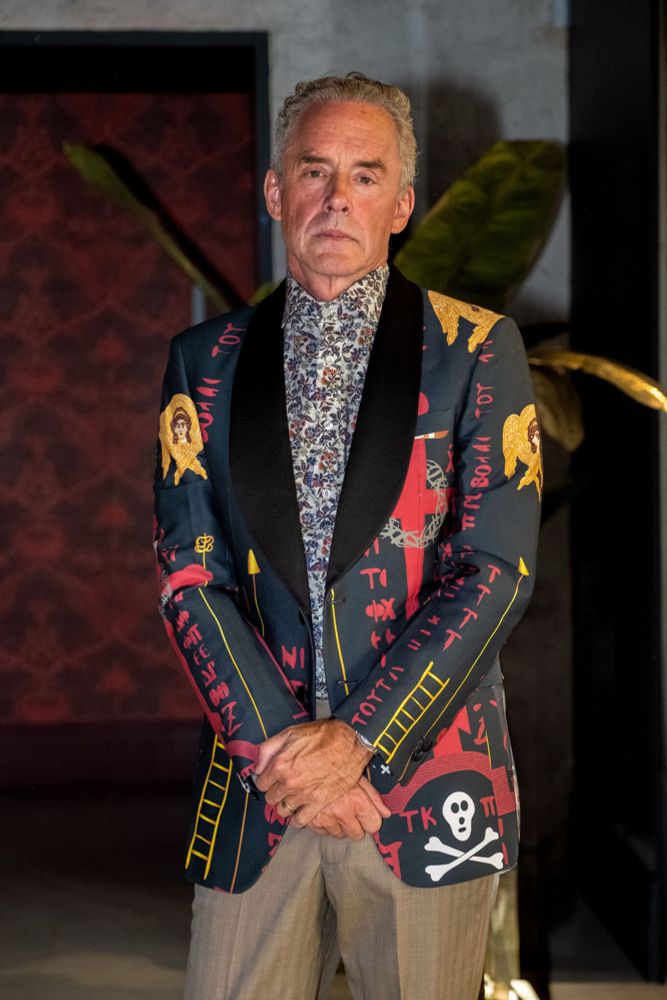 Jordan Pererson poses morosely, hands crossed, in yet another one of his weird suits - this one looks like it was made by AI.