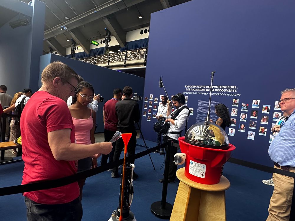 Attendees observe and discuss a deep ARGO float, an oceanographic instrument, on display at an exhibition booth labeled “Explorers of the Deep – Pioneers of Discovery.” A man in a red shirt explains the device to amina schartup’s daughter-sonia, while others nearby film or engage with the exhibit. A wall behind them displays portraits of featured explorers, including amina :)