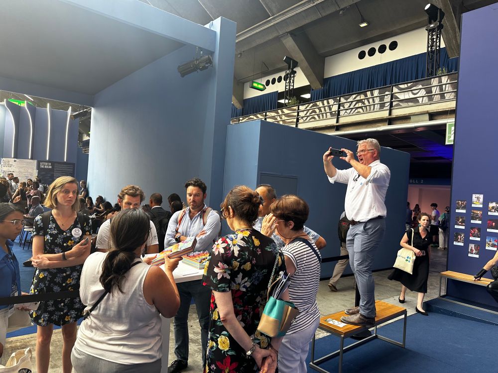 A group of people gathers around a speaker holding colorful 3D printed maps at an indoor event with blue walls and informational posters. One man stands on a bench to take a photo of the group with his phone. The setting is a science/outreach exhibition, with an engaged audience and busy background activity.