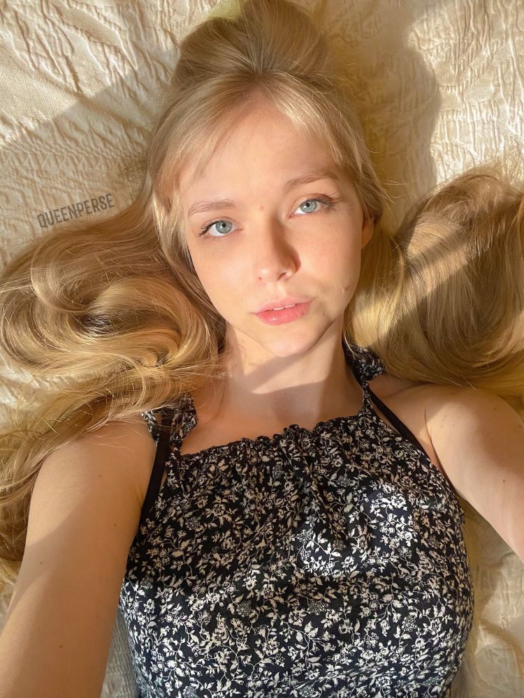 Queen Perse is lying on a textured cream-colored surface, with her hair spread around her. She wears a black sleeveless top with a white floral pattern. Sunlight casts a warm glow across her face, eyes and hair.