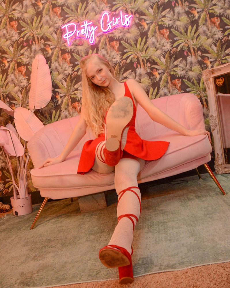 Queen Perse poses playfully on a pink velvet couch, wearing a red dress and red heels. One leg is raised toward the camera, showing the sole of her shoe. Behind her is a neon pink sign reading “Pretty Girls” and a wallpaper with a tropical leaf and animal print. The room has soft pink lighting, a large mirror, and decorative plants, creating a whimsical, feminine atmosphere.