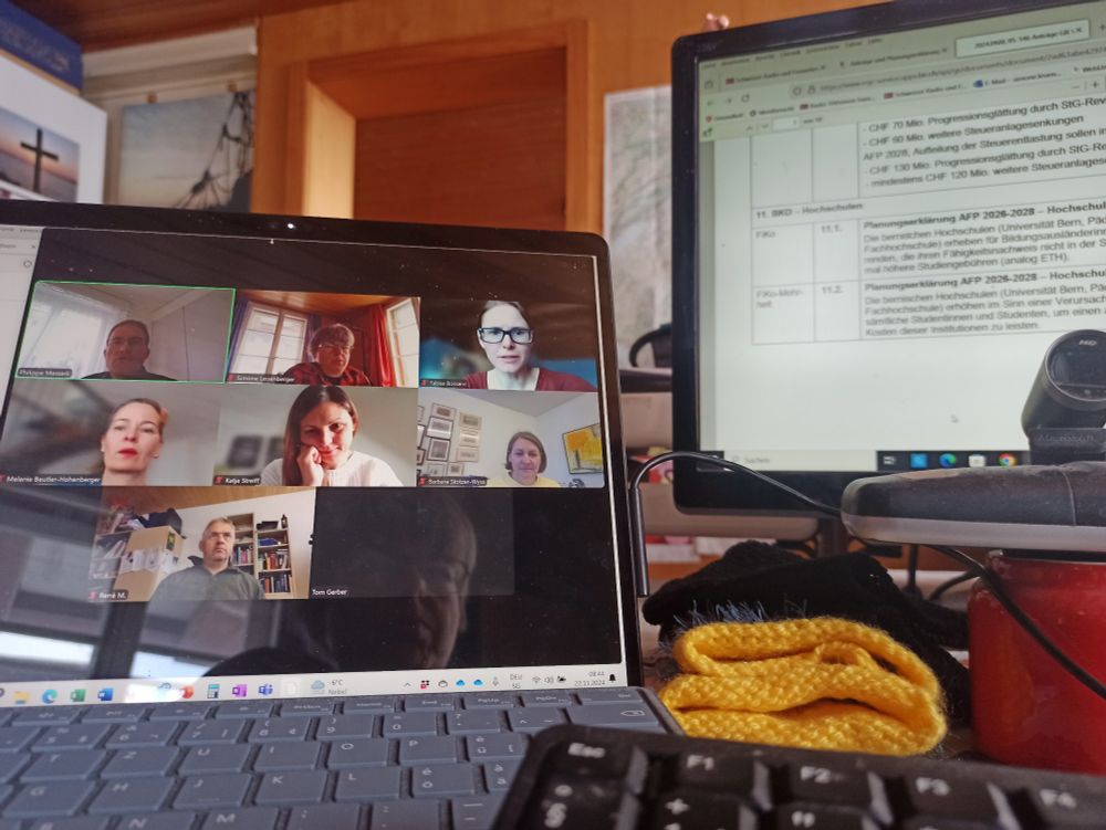 Zwei Bildschirme. Auf dem einen Köpfe eines Online-Meetings, auf dem anderen Text. Im Hintergrund Zimmer Atmosphäre.
