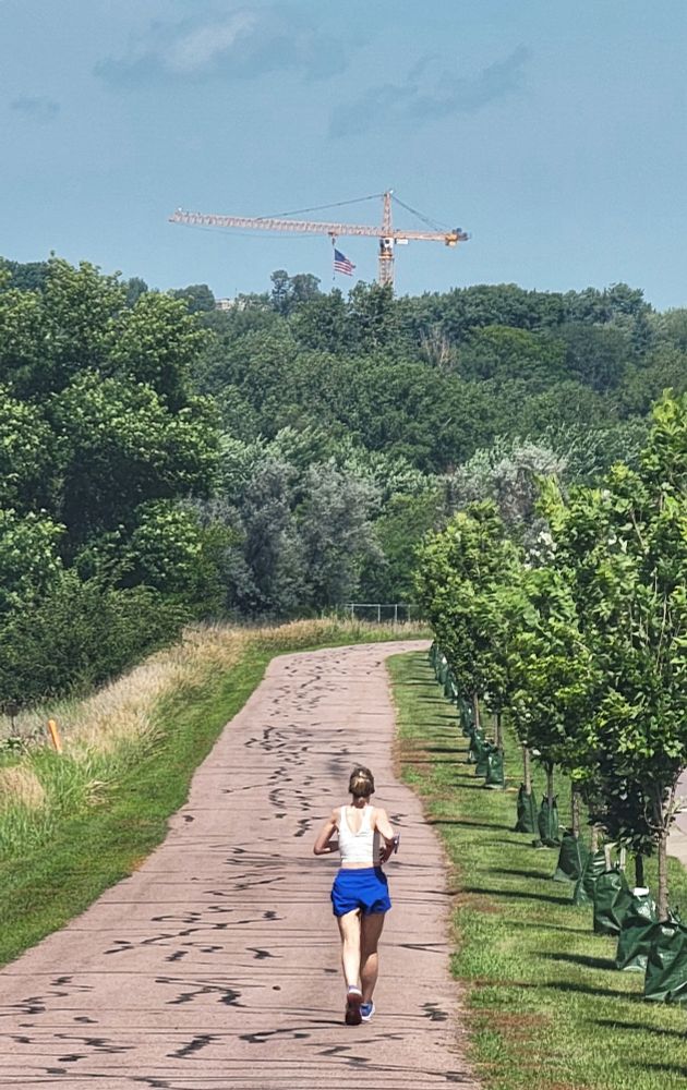 A woman running on a paved trail, lined with newly planted trees. In the distance, the American flag hangs from a crane, just visible over a grove of trees.