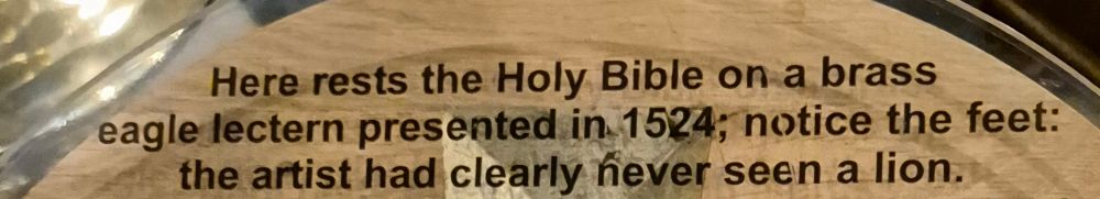 A photograph of part of some Church decoration interpretive text.  It reads:  "Here rests the Holy Bible on.a brass eagle lectern presented in 1524; notice the feet: the artist had clearly never seen a lion."