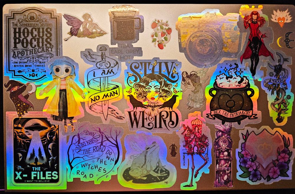 A rose gold colored laptop covered in holographic stickers such as "stay weird," "hex the patriarchy," "hocus pocus apothecary" 