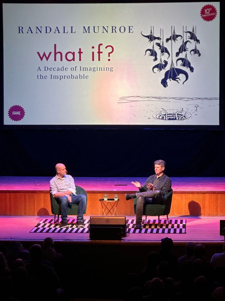 Randall Munroe talking with Matt Parker on a stage with the “What If?” poster projected on the backdrop behind them.