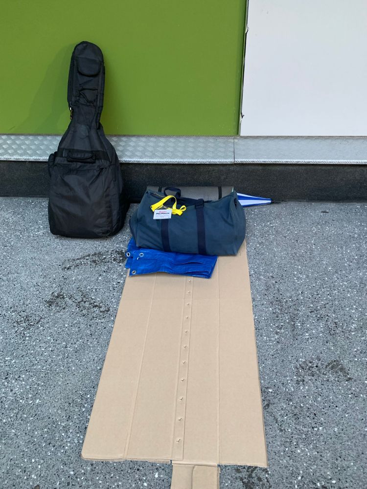 A piece of cardboard in front of a wall. On the cardboard a blue bag and a tarpaulin can be seen. A guitar case is on the left. 