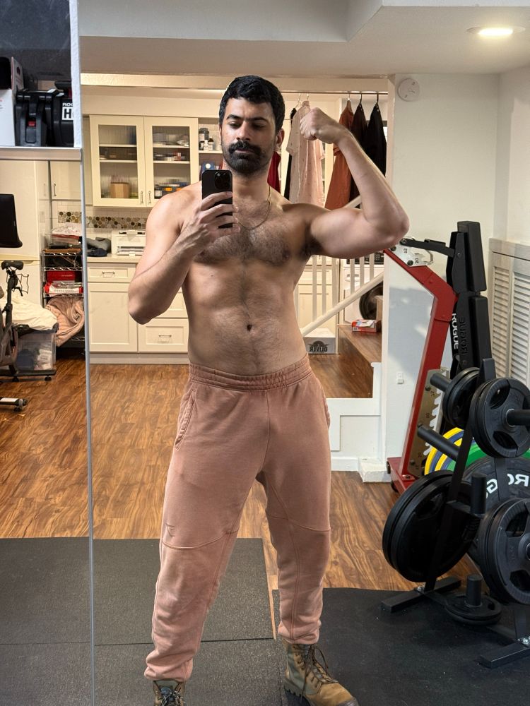 shirtless mirror pic in the home gym wearing washed out red sweatpants and brown boots. flexing my right bicep. 