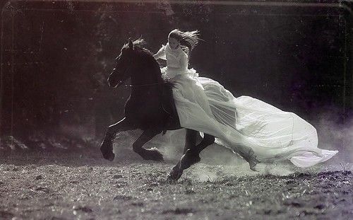 Black and white photograph of woman in long flowing dress atop a galloping horse 
