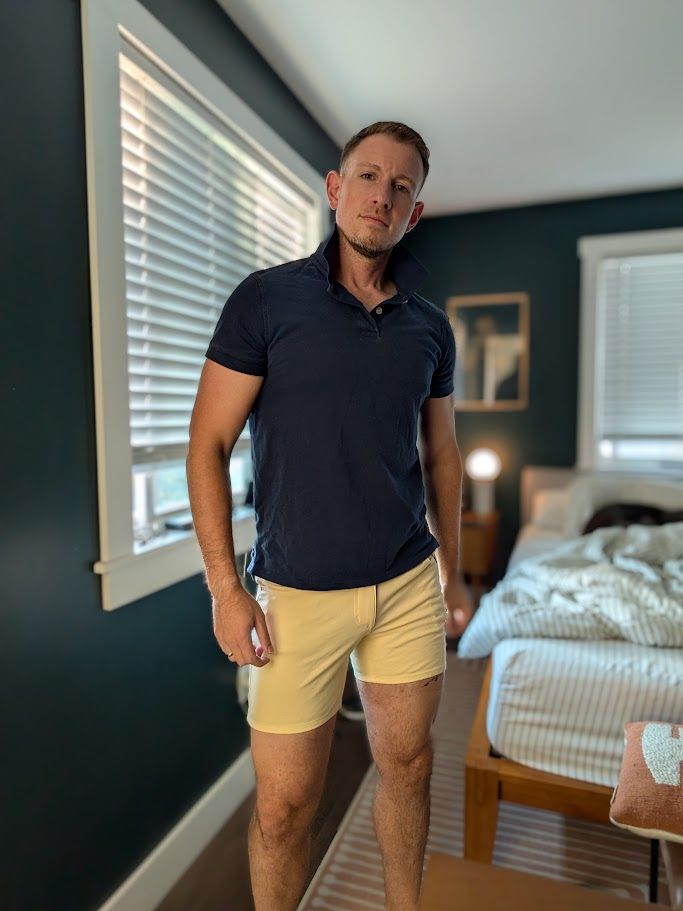 Sam stands in his bedroom wearing a navy blue polo shirt, and very short yellow short. His new thigh tattoo slightly peaks below the shorts on the left leg.