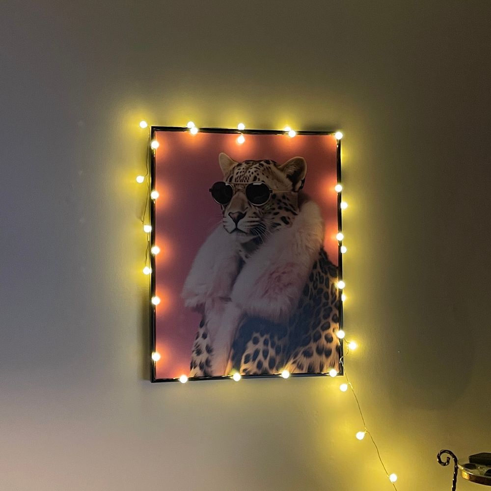 A leopard art print, the leopard is wearing a pink fur-lined coat and sunglasses against a bright pink background. Surrounding the art print are white ball lights. It is fabulously tacky. 