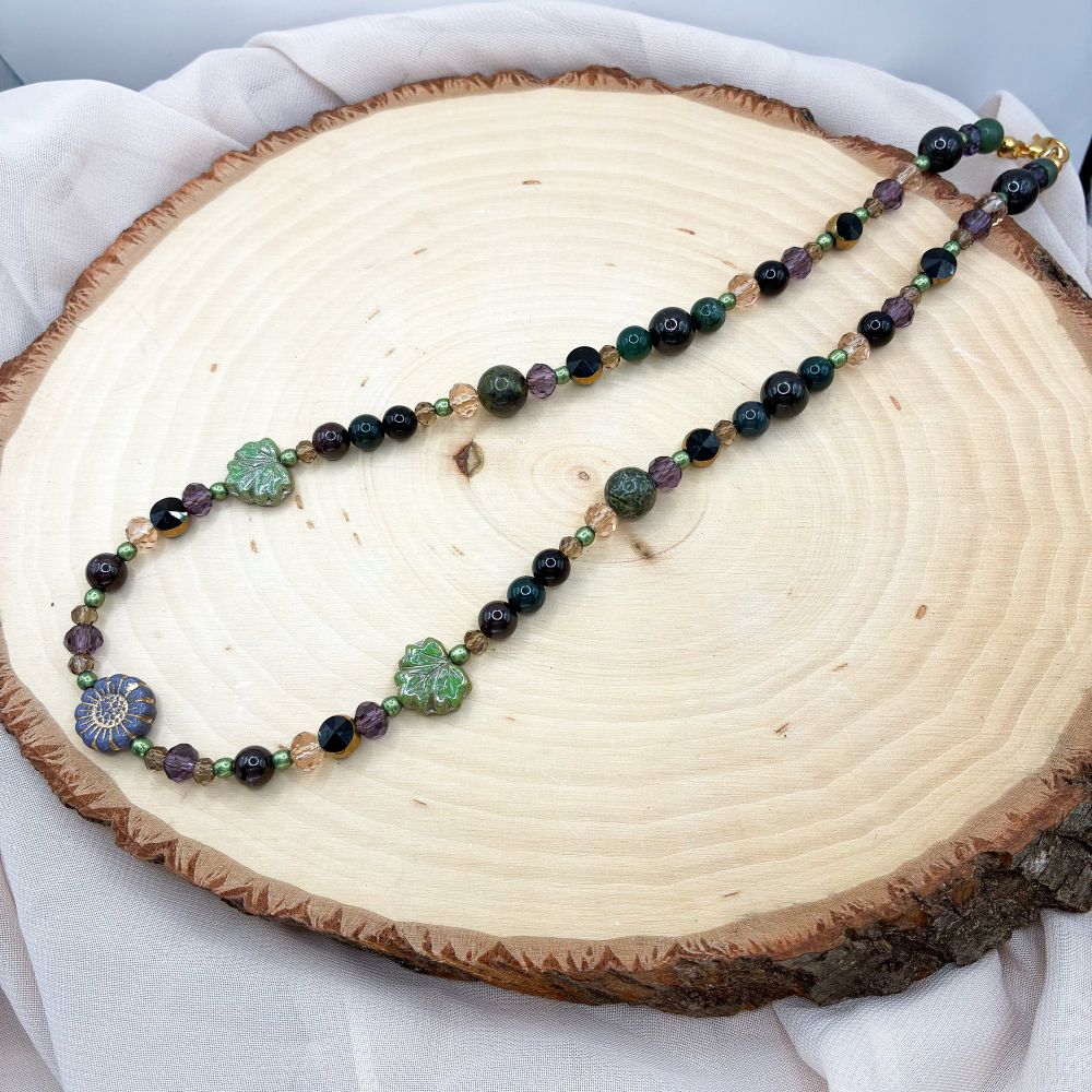 A beaded necklace with a purple flower bead, green leaf beads, and a variety of purple, black, tan, and green beads 