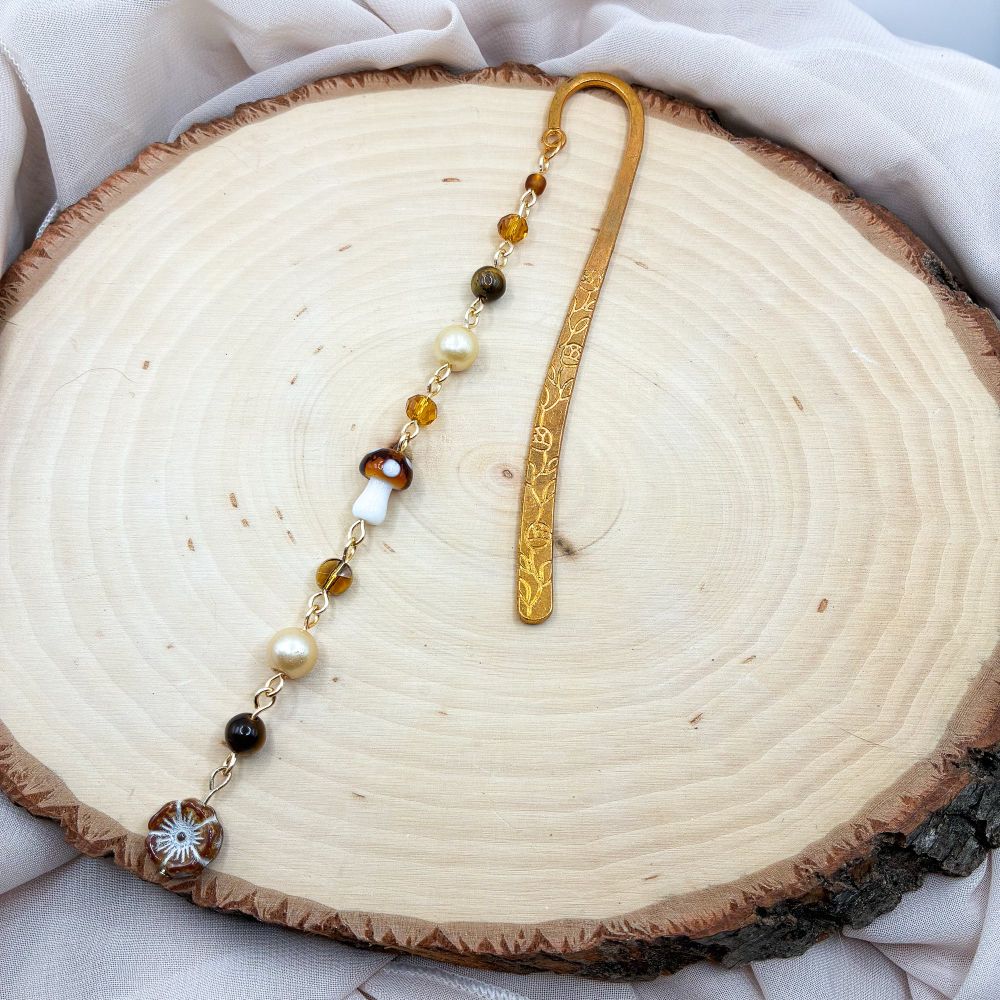 A gold bookmark with a variety of earth tone beads. There is a mushroom bead in the middle and a brown flower bead at the bottom 