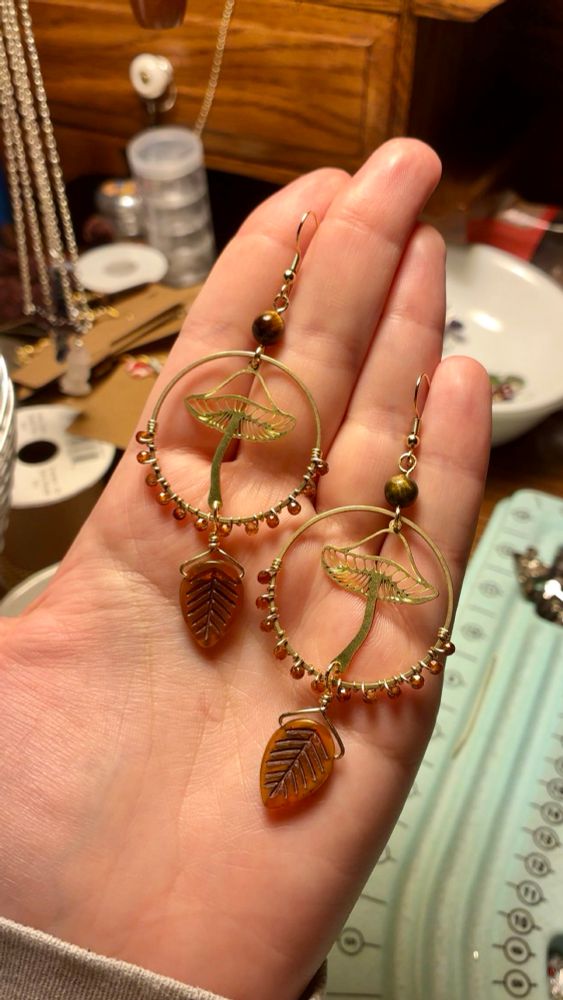 A pair of earrings I made. I wire wrapped small orange hessonite garnet beads onto a hoop and dangled brown leaf beads from the bottom. Inside the hoops are gold mushroom charms 