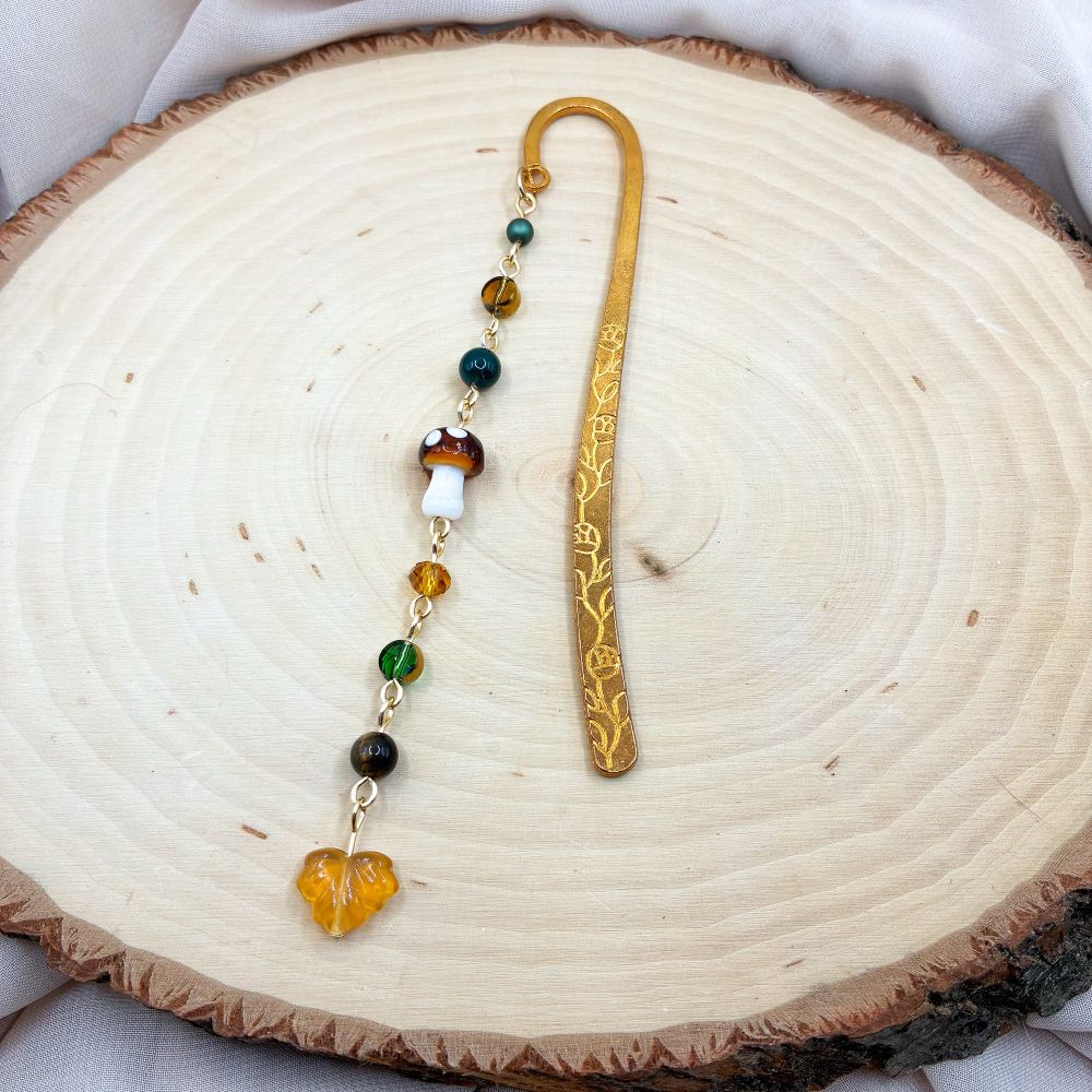 A gold bookmark with a variety of earth tone beads. There is a mushroom bead in the middle, and a yellow leaf bead dangling from the bottom 