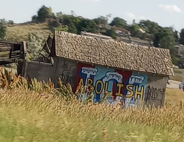 Decorative graffiti on the side of an abandoned roadside building reading "ABOLISH ICE"