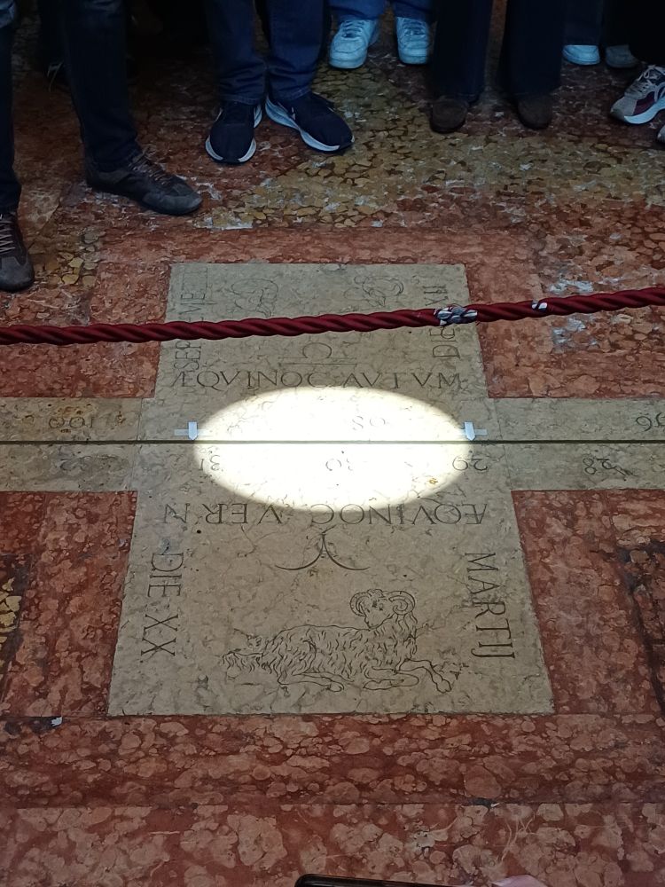 A circle of sunlight marking the solar midday of the vernal equinox upon a sundial. The sundial is a thin line of brass, and the months/signs are engraved upon a slab of white marble. The sun marks both Aries and Libra, being the signs of the two equinoxes. This sundial is the longest in Italy, and can be found in the Basilica of San Petronio in Bologna