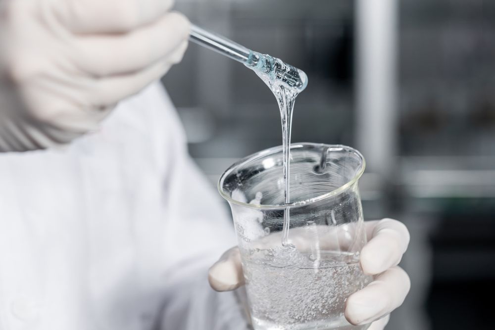 A gloved hand holds a beaker with clear liquid and a dropper in a lab.