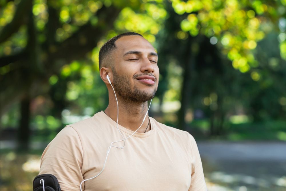 A man in a park, wearing earphones and a light beige t-shirt, with his eyes closed and a peaceful smile on his face.