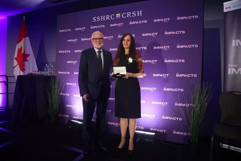 Myriam Denov stands onstage beside a man in a suit as she receives the SSHRC Gold Medal. Denov holds the medal in her hands and smiles for the camera. They are positioned in front of a purple backdrop with the SSHRC/CRSH “Impacts” branding, with a Canadian flag and small table visible to the left.