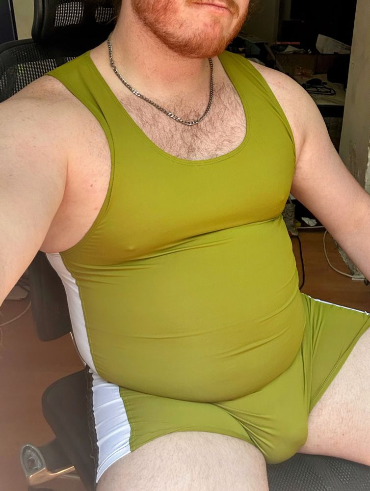 Ginger bear cub wears a very green and very gay wrestling singlet while sitting in an office chair and proudly showing their belly and bulge on tummy Tuesday 