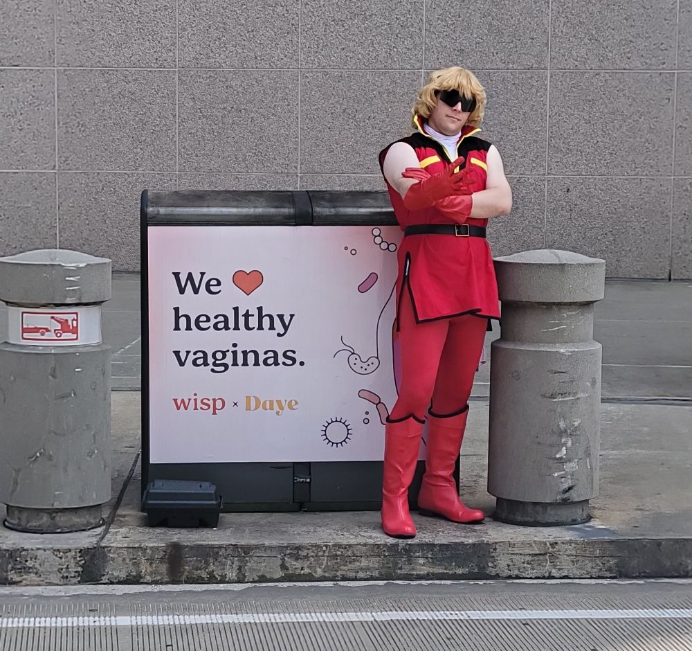 Man cosplaying Quattro Bajeena next to an advertisement that says We ❤️ healthy vaginas.