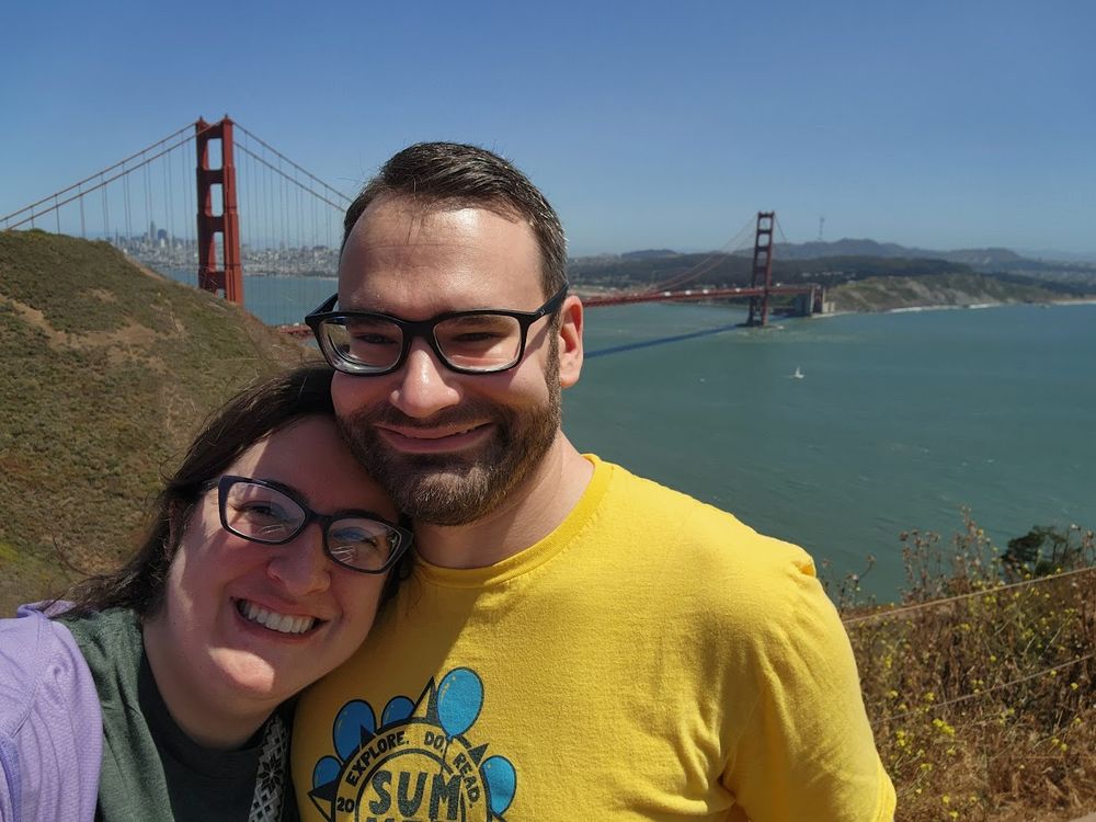 Two people posing in front of the Golden Gate Bridge wearing the CMPLD 2025 Summer Reading t-shirt.