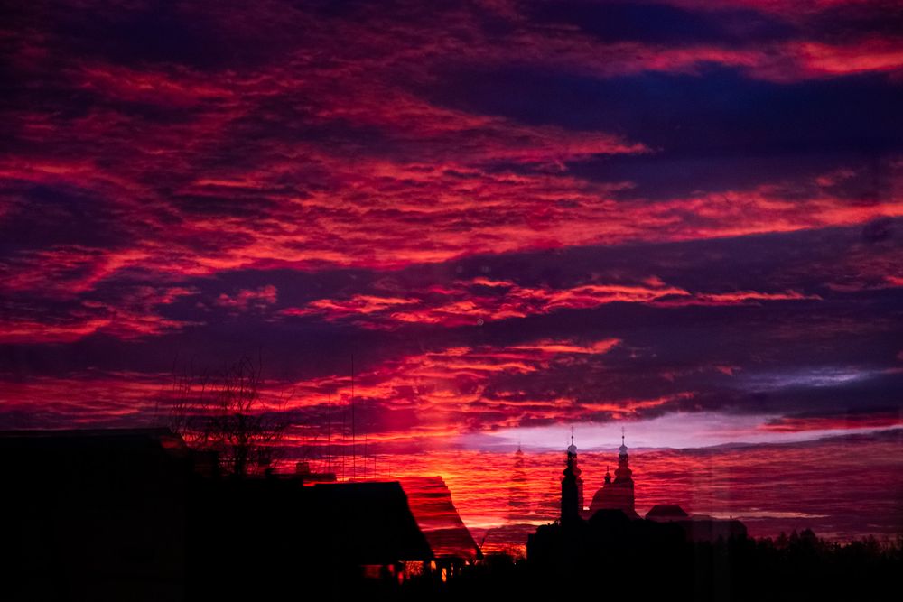 Ein dichtes Wolkenmeer, das von der untergehenden Sonne von unten beleuchtet wird. Stellenweise bereits komplett dunkel, wird es an anderen Stellen in sattem Rot erleuchtet. Im unteren Teil befindet sich die Silhouette einer Kirche. Das Foto wurde über ein dreifach-verglastes Fenster aufgenommen, wodurch die Kirche 3mal in unterschiedlichen Sättigungsgraden und versetzt zu sehen ist.