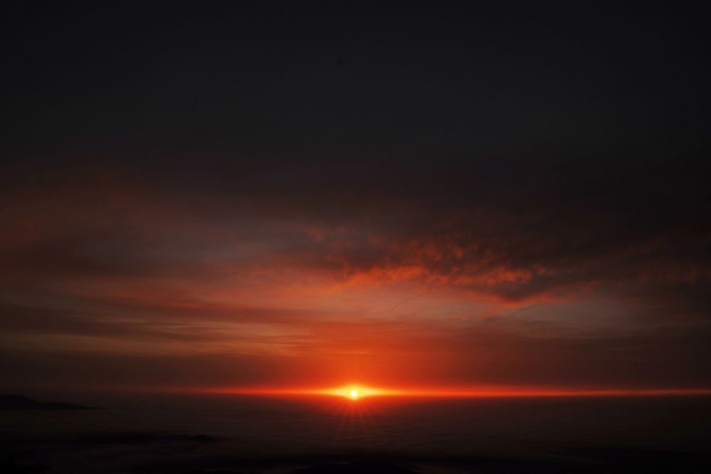 Blick über ein Nebelmeer mit aufgehender Sonne und Wolken. Die Sonne erscheint gerade am Horizont und beleuchtet alles rot.