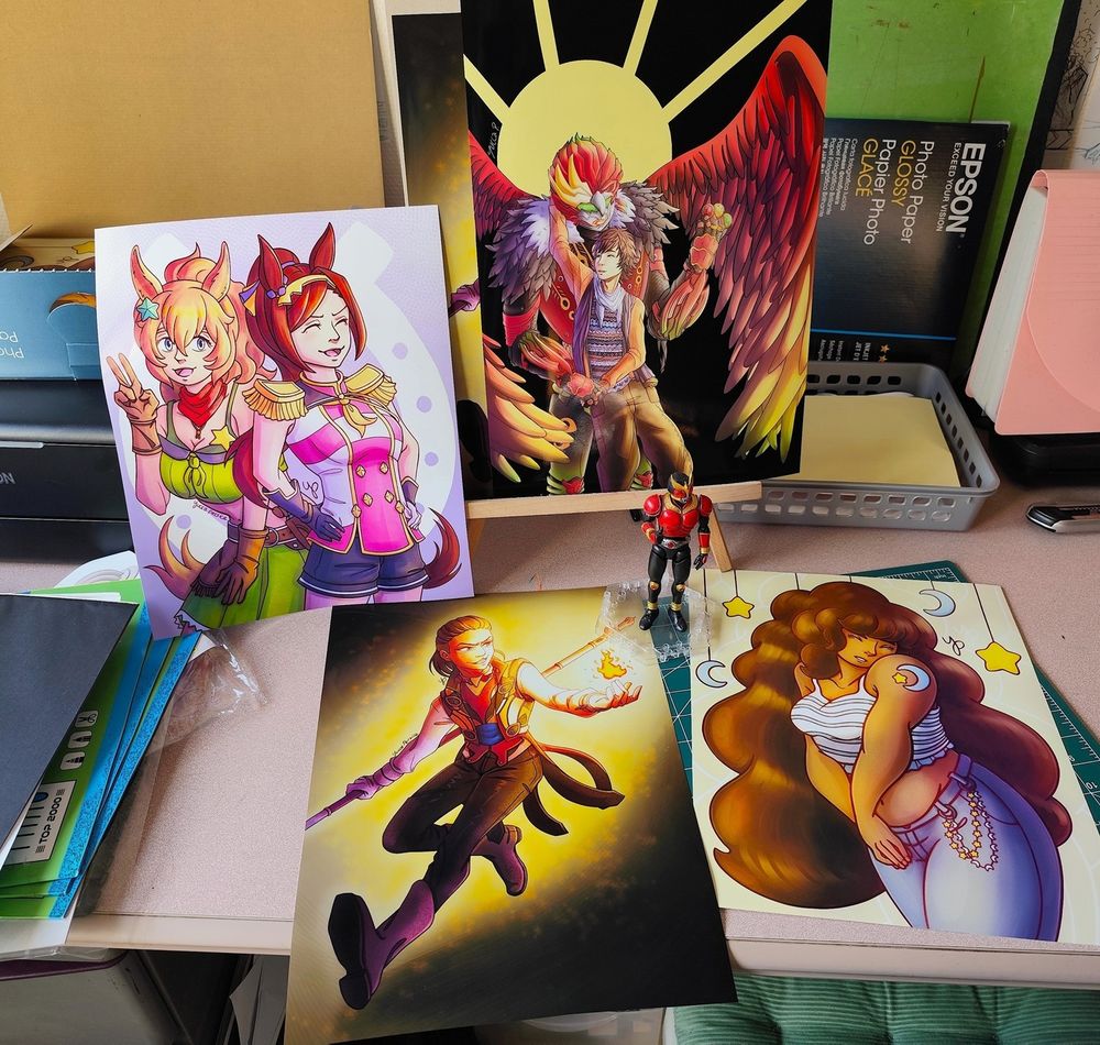 Picture of a few A3 prints of some of my illustrations (Eiji and Ankh from OOO, Sakura Bakushi O and Taiki Shuttle from Umamusume, Fire Cande from Gozyuger, and my OC Luna). There is a Kuuga figure in the middle looking tiny in comparison.