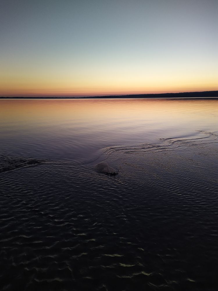 Sonnenuntergang am See. Der Himmel ist wolkenlos, am Horizont sieht man noch die orangerote Färbung nachdem die Sonne untergegangen ist. Der See ist glatt, nur im Vordergrund kräuselt sich das Wasser durch einen Bacheinlauf.