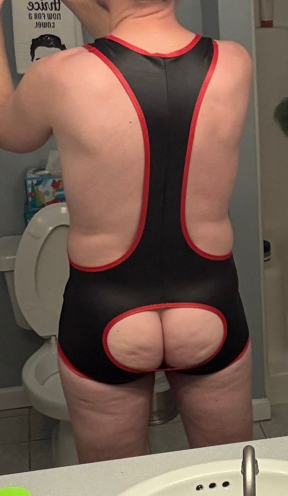 A white man wearing a black singlet with red trim and a cut out in the seat, exposing his butt cheeks. 