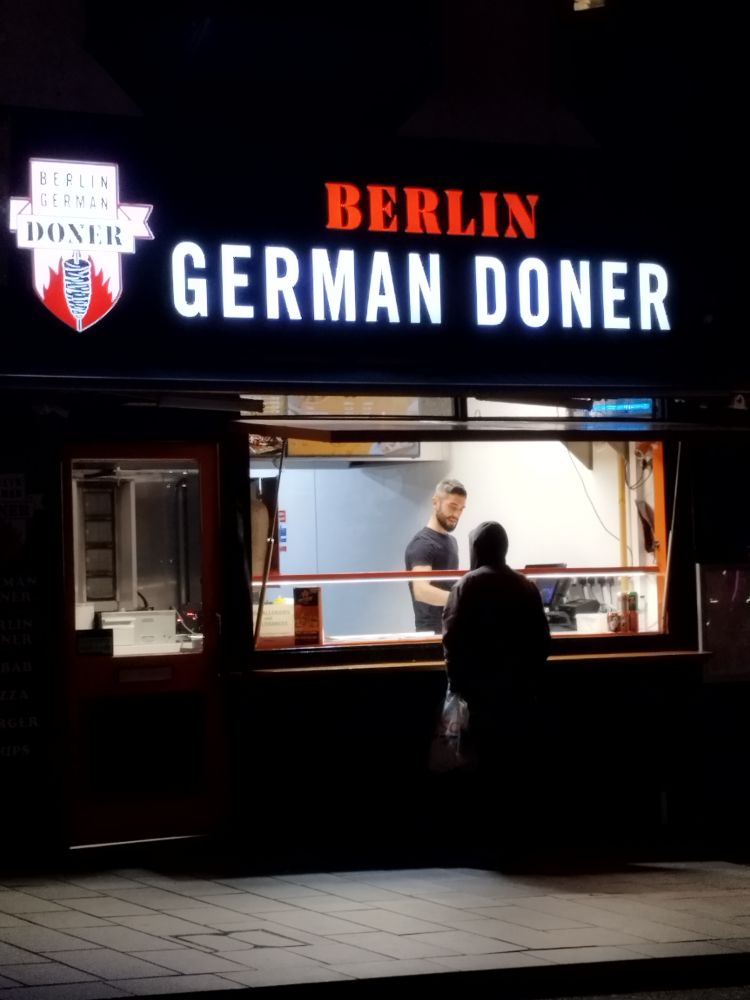 Ein Straßenimbiss mit einem Verkäufer und einer Person vor dem Tresen, welche vermutlich grad Essen bestellt.

Darüber eine große Leuchtschrift 
BERLIN 
German Döner 